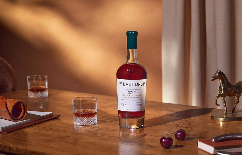 Load image into Gallery viewer, A bottle of The Last Drop Release No. 37 Buffalo Trace 27 Year Old Kentucky Straight Bourbon Whiskey 700ml, a rare expression from Buffalo Trace, sits elegantly on a wooden table alongside two glasses of whiskey, a pair of cherries, a notebook, a pen holder, and a small horse statue in the background—evoking the rich heritage synonymous with Buffalo Trace Distillery.
