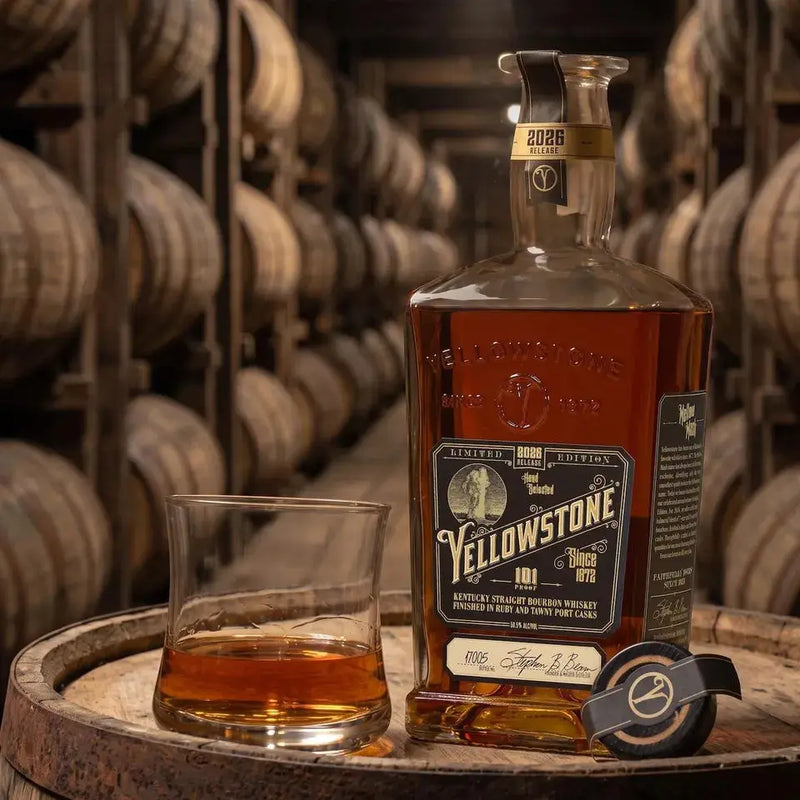 Load image into Gallery viewer, A bottle of Yellowstone 2026 Limited Edition Bourbon Finished in Ruby and Tawny Port Casks, a glass of Kentucky straight bourbon, and a cap sit on a barrel in a dimly lit aging room lined with wooden barrels.
