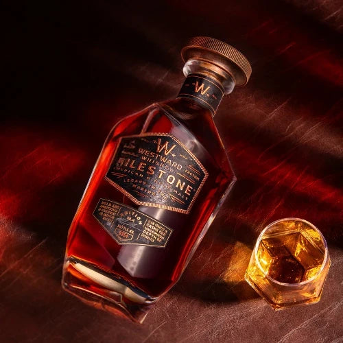 Load image into Gallery viewer, A bottle of Westward Whiskey Milestone Edition No. 4 American Single Malt (Pre-Order) rests on a brown surface beside a glass filled with the same whiskey, both highlighted by warm, angled lighting.

