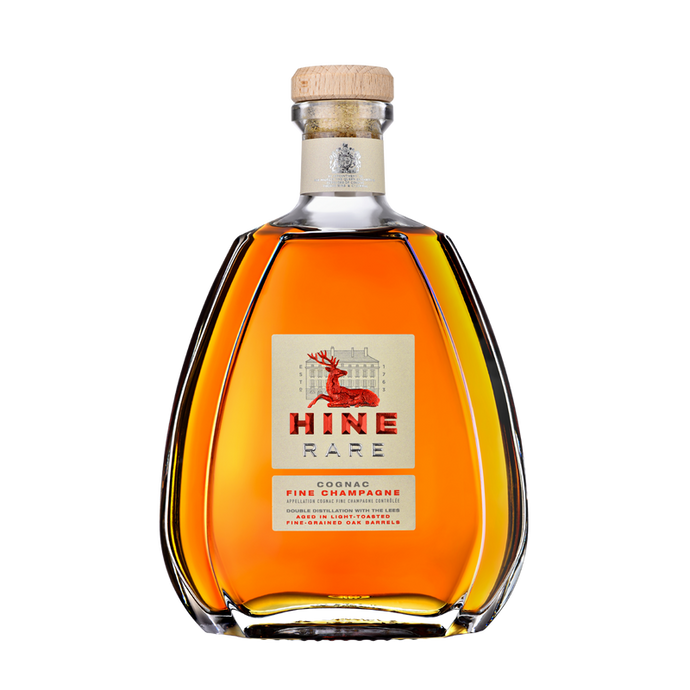A clear, curvy bottle of HINE Cognac Rare by HINE Cognac features a wooden cork and a label with a red deer and gold accents. The amber liquid shows through the glass, all set against a plain white background.