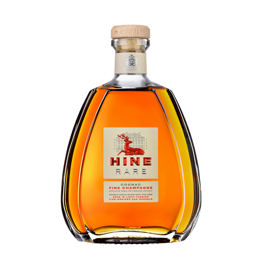 A clear, curvy bottle of HINE Cognac Rare by HINE Cognac features a wooden cork and a label with a red deer and gold accents. The amber liquid shows through the glass, all set against a plain white background.