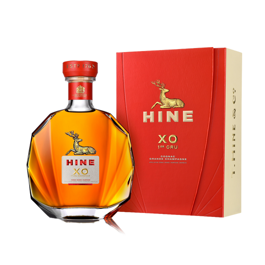 A bottle of HINE XO 1er Cru Cognac by HINE Cognac with a gold deer logo and red label stands beside its matching red and gold box, both featuring the same design, all set against a white background.