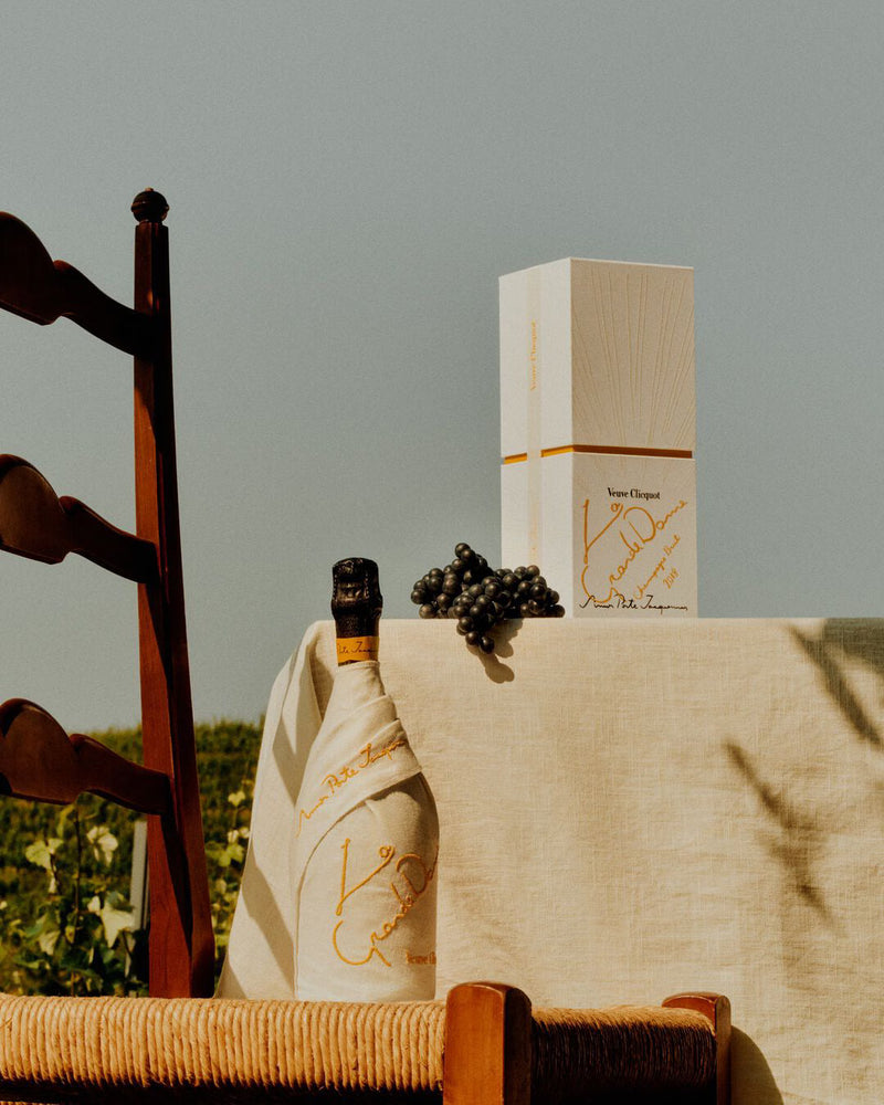 Load image into Gallery viewer, A wooden chair stands by a table with a light cloth, displaying Veuve Clicquot La Grande Dame 2018 Limited Edition by Simon Porte Jacquemus, grapes, and a white gift box outdoors amid lush greenery.
