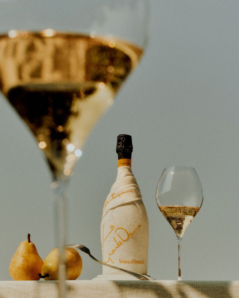 Load image into Gallery viewer, A bottle of Veuve Clicquot La Grande Dame 2018 Limited Edition by Simon Porte Jacquemus, in tissue wrap, sits with two champagne glasses and two pears on a white surface against a soft, neutral backdrop.
