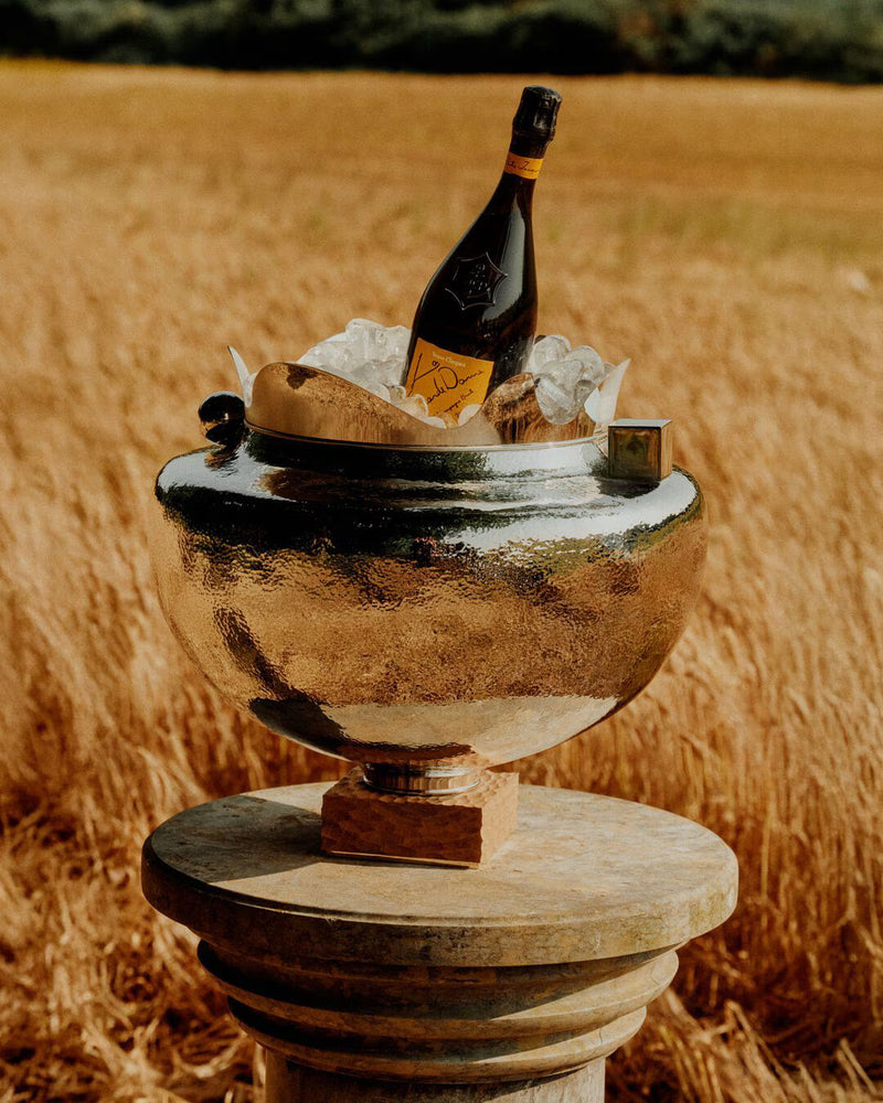 Load image into Gallery viewer, A bottle of Veuve Clicquot La Grande Dame 2018 Limited Edition by Simon Porte Jacquemus chills in a silver ice bucket on a stone pedestal amid golden wheat—an exclusive Veuve Clicquot champagne moment.
