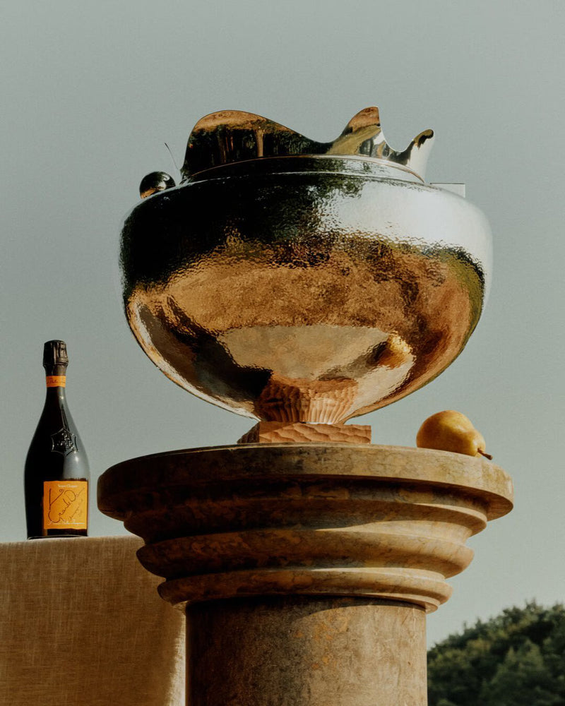 Load image into Gallery viewer, A golden bowl on a stone pedestal sits beside a lemon, with Veuve Clicquot La Grande Dame 2018 Limited Edition by Simon Porte Jacquemus in the background, all set outdoors beneath a clear sky.
