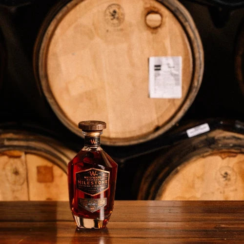 Load image into Gallery viewer, A bottle of Westward Whiskey Milestone Edition No. 4 American Single Malt (Pre-Order) from Westward rests on a wooden surface before towering barrels, softly illuminated to create a warm, inviting atmosphere that showcases the craftsmanship behind this exceptional whiskey.
