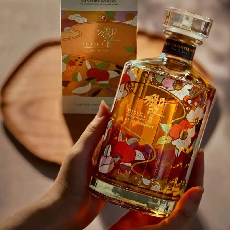 Load image into Gallery viewer, Hands holding a Hibiki Japanese Harmony Chiso 2026 Limited Edition Blended Japanese Whisky 750mL bottle decorated with colorful floral illustrations, with its matching gift box standing behind it on a wooden board.
