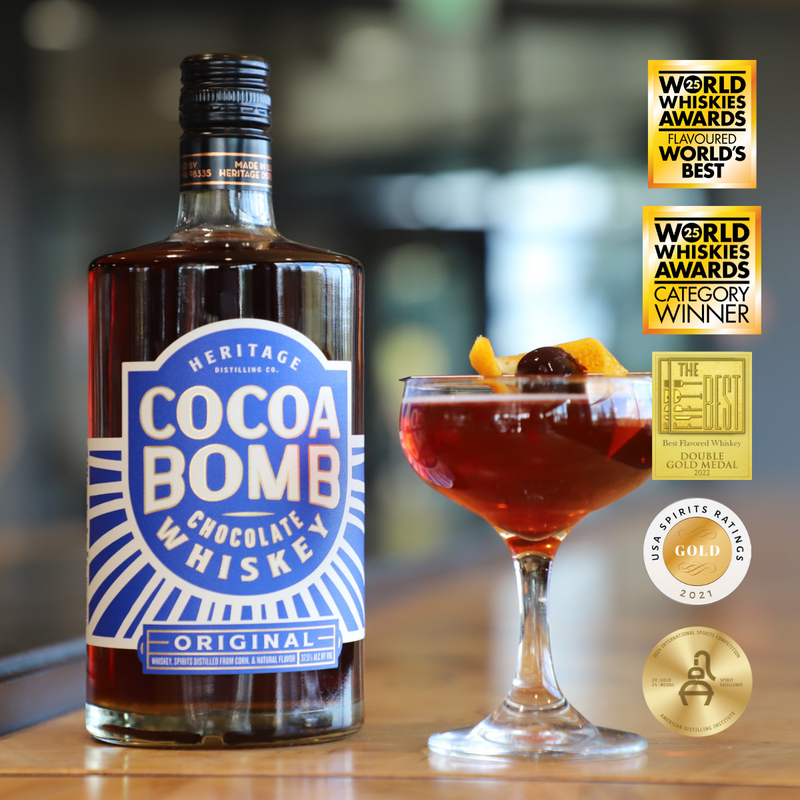 Load image into Gallery viewer, Two bottles of Heritage Distilling Cocoa Bomb Whiskey are placed next to a chocolate whiskey cocktail garnished with an orange twist and cherry, while several award medals and badges are displayed on the right.
