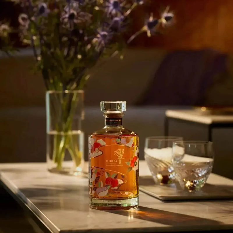 Load image into Gallery viewer, A glass bottle of Hibiki Japanese Harmony Chiso 2026 Limited Edition Blended Japanese Whisky 750mL decorated with colorful floral motifs, sitting on a table next to two empty drinking glasses and a vase of flowers.

