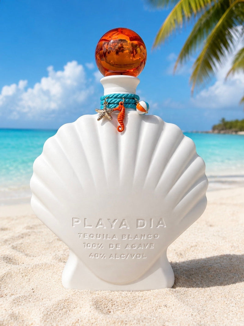 Load image into Gallery viewer, A shell-shaped white bottle of Playa Dia Blanco Tequila rests on a sandy beach with a turquoise sea, palm frond, and blue sky in the background; this 100% Blue Weber agave, additive-free tequila features an orange stopper and is adorned with seashell charms.

