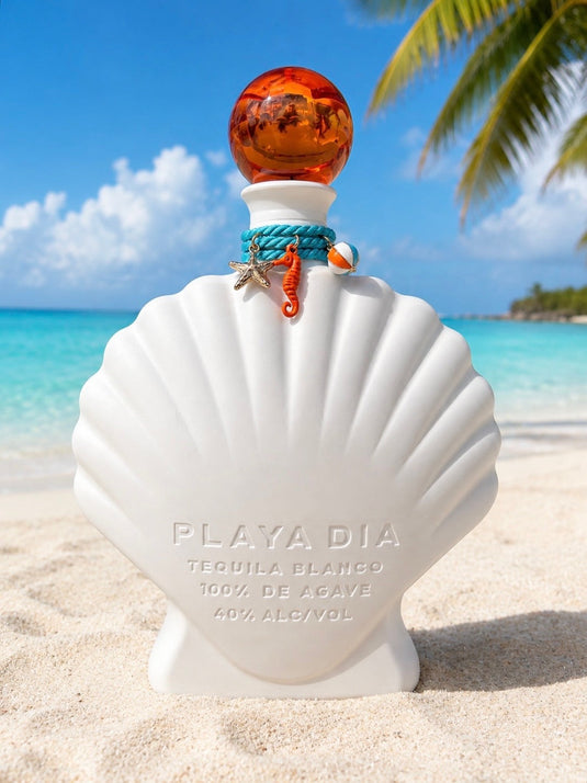 A shell-shaped white bottle of Playa Dia Blanco Tequila rests on a sandy beach with a turquoise sea, palm frond, and blue sky in the background; this 100% Blue Weber agave, additive-free tequila features an orange stopper and is adorned with seashell charms.