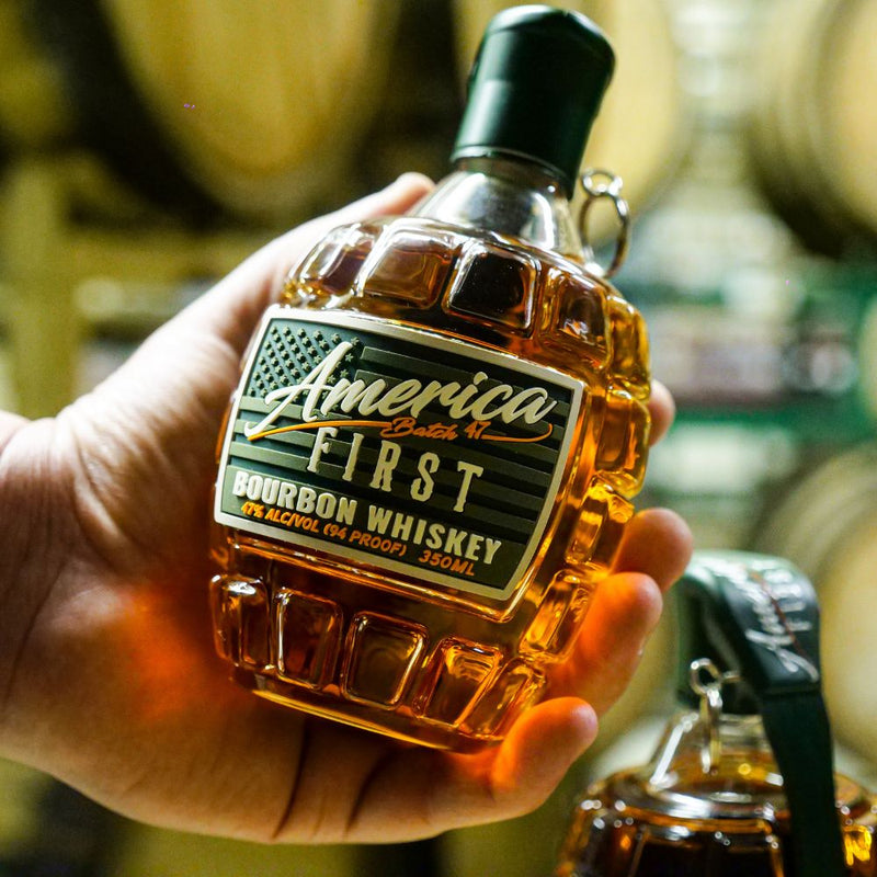 Load image into Gallery viewer, A hand holds an America First Grenade Straight Bourbon Whiskey 2 Pack bottle, shaped like a grenade and labeled 94 proof, 350ml—ideal as a collectible or patriotic gift, with wooden barrels blurred in the background.
