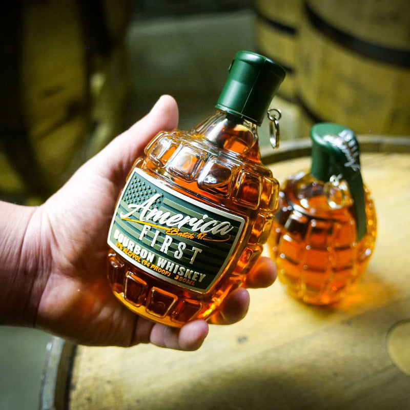 Load image into Gallery viewer, A hand holds an America First Grenade Straight Bourbon Whiskey bottle by America First, shaped like a grenade, with another in the background—ideal for patriotic gifting. Available as a 2 Pack.
