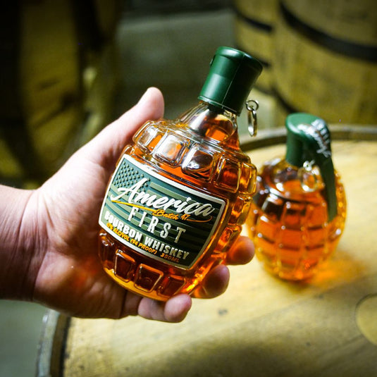 A hand holds an America First Grenade Straight Bourbon Whiskey bottle by America First, shaped like a grenade, with another in the background—ideal for patriotic gifting. Available as a 2 Pack.
