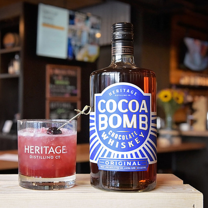 Load image into Gallery viewer, Two bottles of Heritage Distilling Cocoa Bomb Whiskey sit on a wooden surface next to a glass of pink whiskey cocktail garnished with berries in a cozy bar setting.
