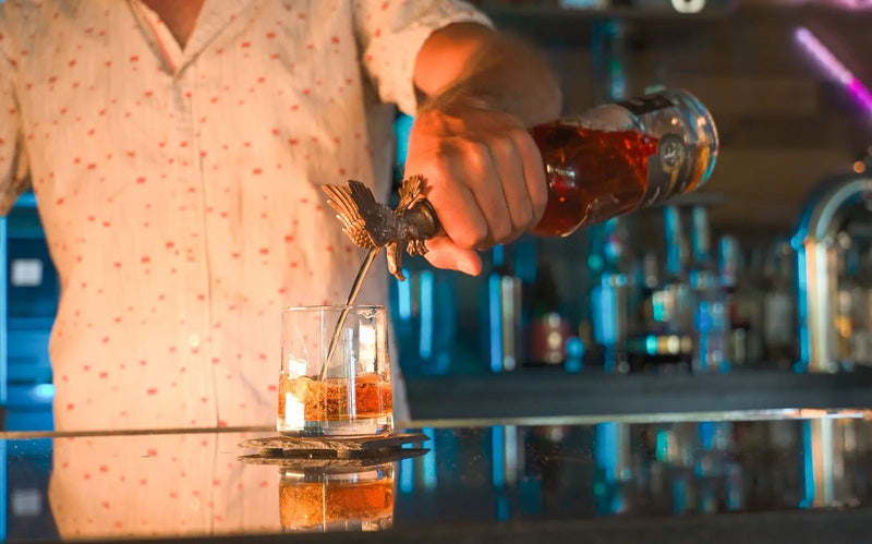 Load image into Gallery viewer, A bartender pours America First 250 Year Limited Edition Bourbon—a special 10-year-aged release—into a glass with ice from a bottle topped with an eagle-shaped stopper, set on a reflective bar counter.
