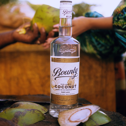A bottle of Bounty Rum Coconut 750 ml stands on a wooden surface surrounded by opened coconuts, while blurred hands holding coconuts appear in the background.
