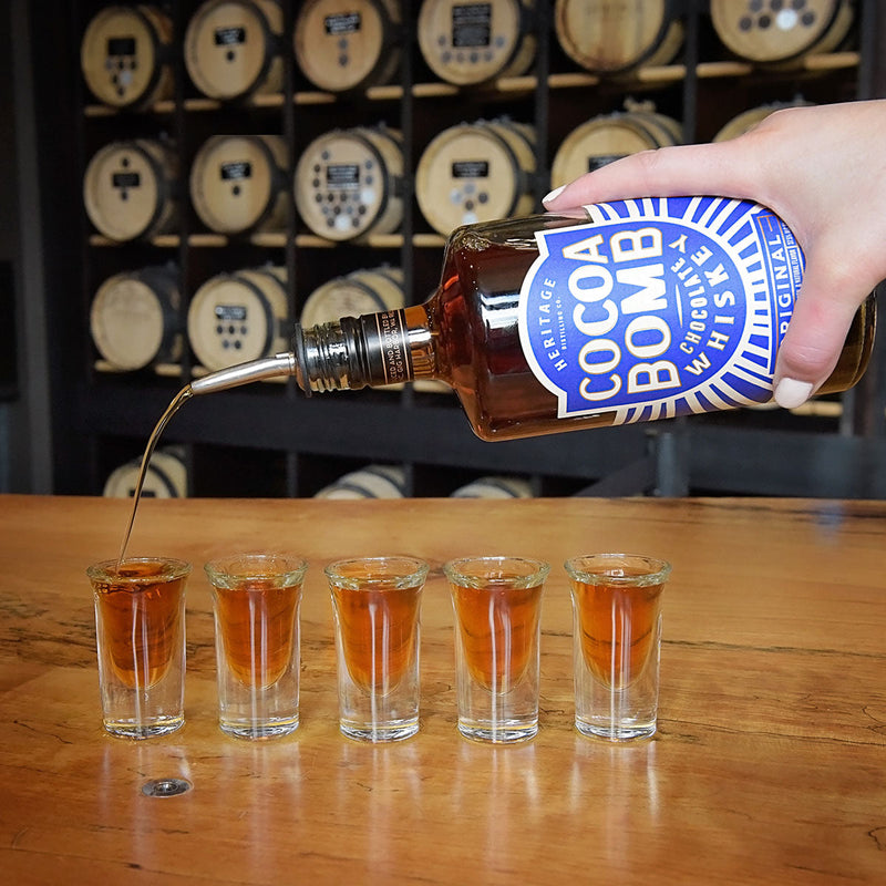 Load image into Gallery viewer, A hand pours Heritage Distilling Cocoa Bomb Whiskey - 2 Bottle into one of five shot glasses lined up on a wooden bar, with whiskey barrels stacked in the background.
