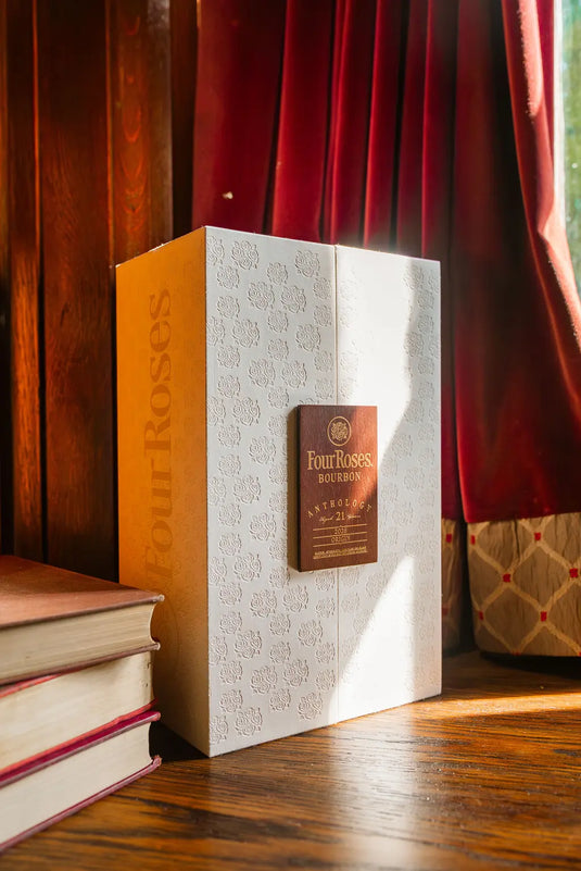 A white embossed box of Main Street Liquor's Four Roses The Anthology Series Chapter 1: Origin 21 Year Bourbon sits on a wooden floor near stacked books, red curtains, and sunlight—a stunning collectible bourbon display.