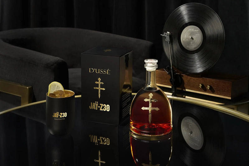 Load image into Gallery viewer, A bottle of D’USSE VSOP Jay-Z30 Limited Edition Cognac (PRE-ORDER) stands in a branded black box beside a glass with a lemon garnish, on a reflective table. A record player and velvet chair in the background add to the French cognac’s luxurious ambiance.
