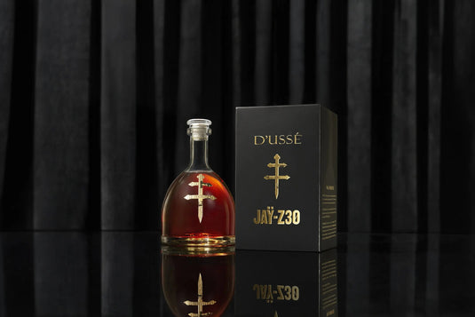 A bottle of D’USSÉ VSOP Jay-Z30 Limited Edition Cognac (PRE-ORDER) stands beside its striking black and gold box, set against a vertically striped black background, showcasing this exclusive collaboration from D’USSÉ.