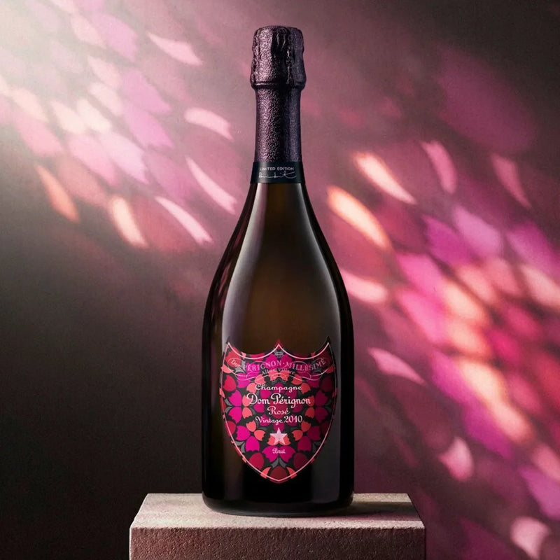 Load image into Gallery viewer, A bottle of Dom Pérignon Rosé Vintage 2010 Damien Hirst Limited Edition Champagne with a decorative, multi-colored floral label stands on a square stone pedestal against a pink and purple patterned background.
