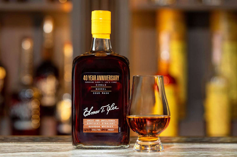 Load image into Gallery viewer, A bottle of Elmer T. Lee 40 Year Anniversary Uncut &amp; Unfiltered Single Barrel Bourbon 127.8 Proof sits beside a whiskey glass of amber liquid on a wooden surface, with blurred bottles in the background.
