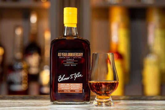 A bottle of Elmer T. Lee 40 Year Anniversary Uncut & Unfiltered Single Barrel Bourbon 127.8 Proof sits beside a whiskey glass of amber liquid on a wooden surface, with blurred bottles in the background.
