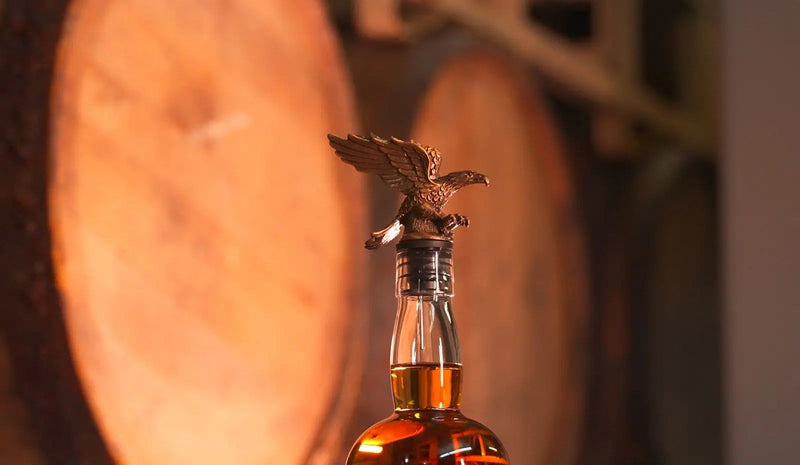 Load image into Gallery viewer, A close-up of the America First 250 Year Limited Edition Bourbon bottle by America First, featuring a bronze eagle stopper, with large wooden barrels blurred in the background.
