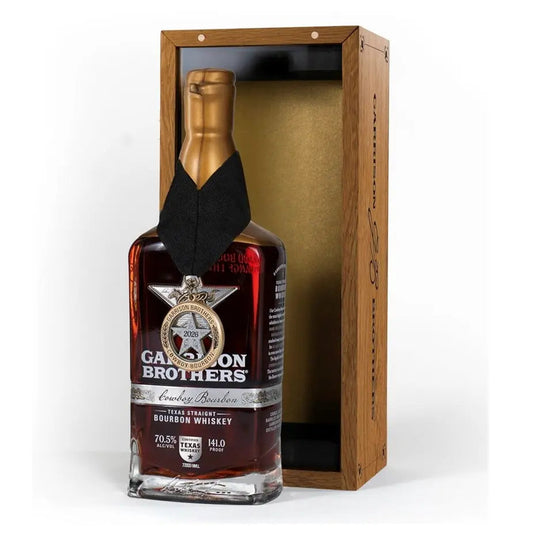 A bottle of Garrison Brothers 2026 Cowboy Bourbon Texas Straight Bourbon Whiskey, proudly bearing a medal around its neck, is showcased beside an open wooden box featuring a gold interior.