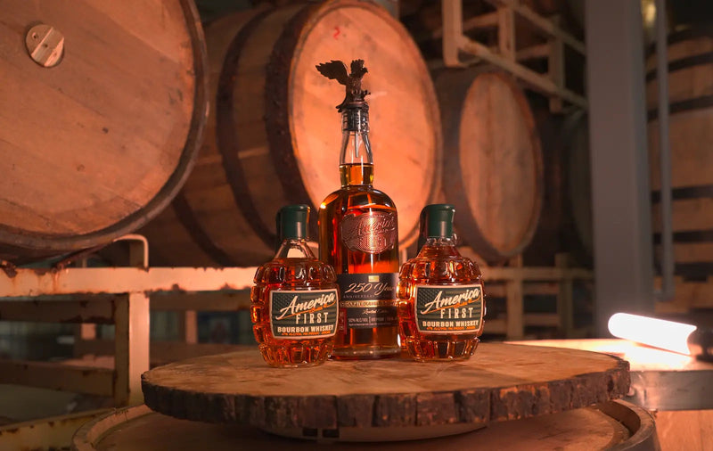 Load image into Gallery viewer, Three bottles from the America First Limited Edition Bourbon Bundle—featuring the Anniversary Bottle and two Grenade Whiskeys—are displayed on a wooden barrel, with large aging barrels in the background, creating a warm distillery setting reminiscent of classic America First Grenade Whiskey Bottles.
