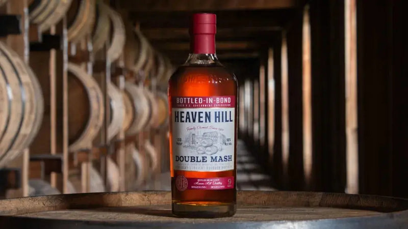 Load image into Gallery viewer, A bottle of Heaven Hill Double Mash Bottled-in-Bond Bourbon (Pre-Order) rests on a barrel inside a dimly lit rickhouse, with rows of wooden barrels stacked in the background.

