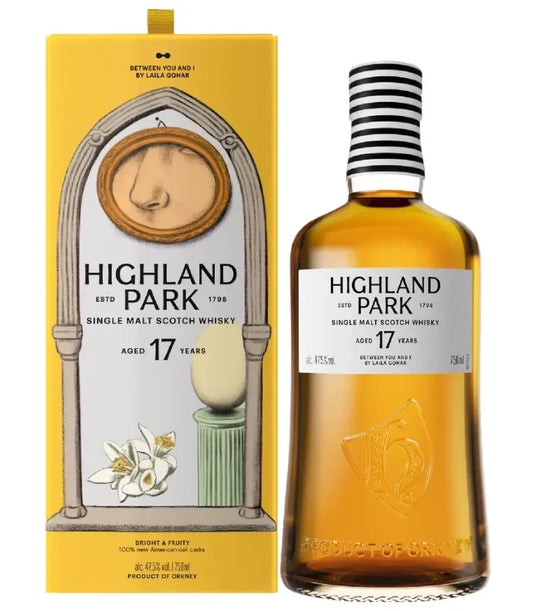 A bottle of Highland Park 17 Year Between You & I Single Malt Scotch Whisky stands next to its yellow box, decorated with artistic images of a nose, flowers, and a ceramic object.