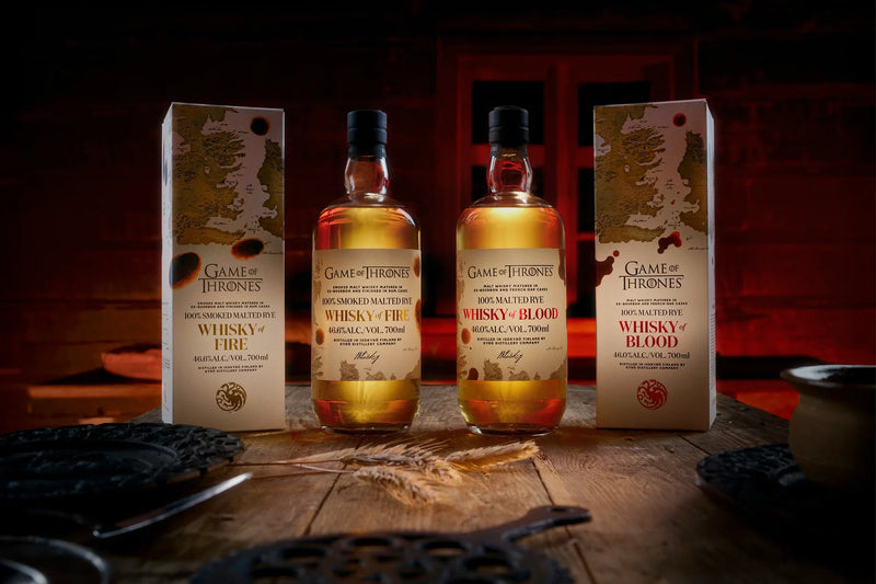 Load image into Gallery viewer, Three bottles of the Kyrö x Game of Thrones Whisky of Fire &amp; Blood Bundle (PRE-ORDER) from Main Street Liquor stand between matching boxes on a rustic wooden table, set against a dramatic, red-lit backdrop.
