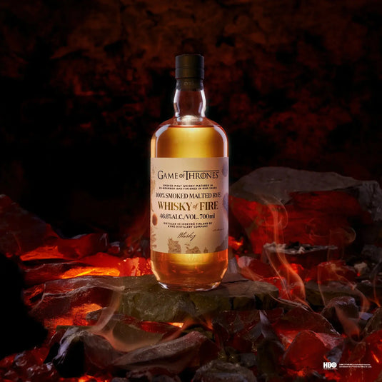 A bottle of Kyrö x Game of Thrones Whisky of Fire & Blood Bundle from Main Street Liquor stands on glowing coals amid swirling smoke, with a fiery backdrop—an epic tribute to Finnish rye whisky craftsmanship. Pre-order now!.