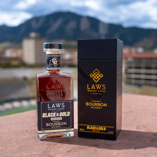 A bottle of Laws Whiskey House Black & Gold Select Four Grain Straight Bourbon Whiskey stands beside its elegant black and gold box from Laws Whiskey House, with a blurred cityscape and mountain backdrop.