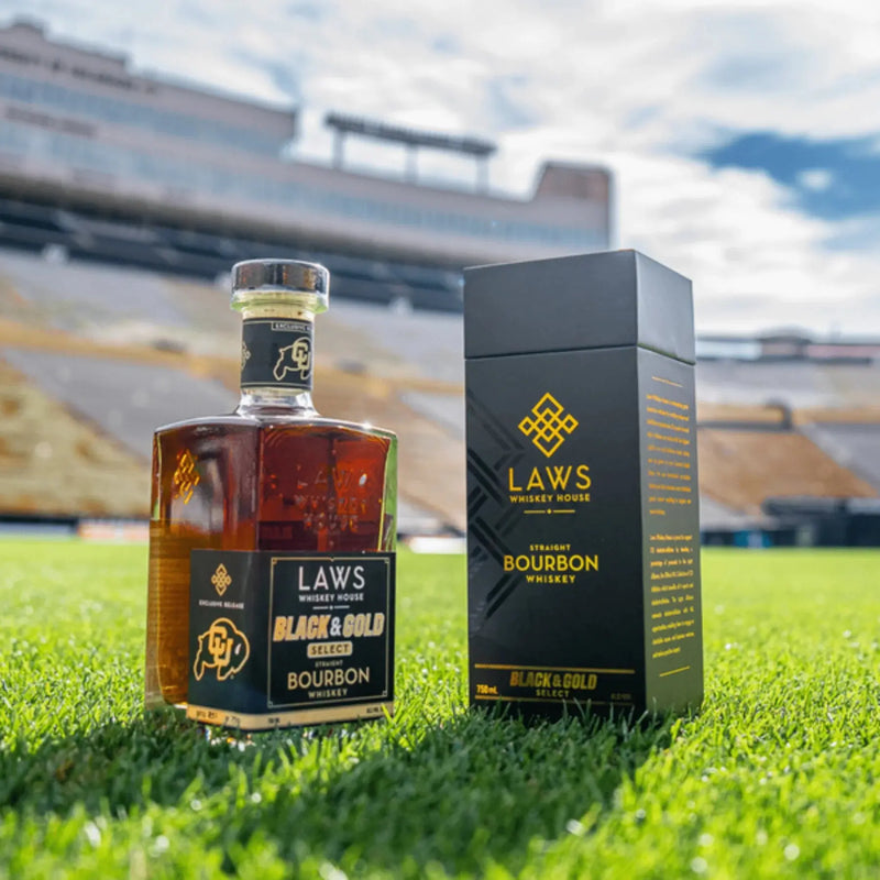 Load image into Gallery viewer, A bottle and box of Laws Whiskey House Black &amp; Gold Select Four Grain Straight Bourbon Whiskey rest on lush green grass, with a stadium and bleachers softly blurred in the background beneath a blue sky—showcasing this limited-release whiskey from Laws Whiskey House, crafted with their signature Colorado Four Grain Bourbon.
