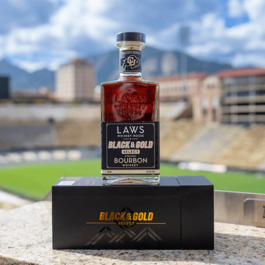 A bottle of Laws Whiskey House Black & Gold Select Four Grain Straight Bourbon Whiskey from Laws Whiskey House, a limited-release expression, rests on its box against the backdrop of a blurred stadium and the majestic Colorado mountains beneath a partly cloudy sky.