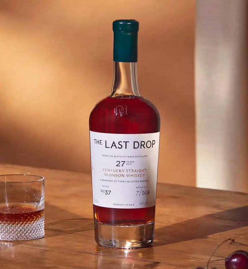 Load image into Gallery viewer, A bottle of The Last Drop Release No. 37 Buffalo Trace 27 Year Old Kentucky Straight Bourbon Whiskey 700ml, crafted by the acclaimed Buffalo Trace brand, stands on a wooden surface next to a glass of whiskey with ice, both illuminated by warm sunlight.
