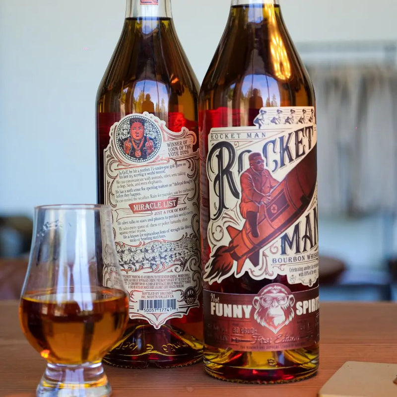 Load image into Gallery viewer, Two bottles of Rocket Man Bourbon Whiskey by Rocket Man are displayed—one featuring the front label with a man riding a rocket, the other the back. A glass of this limited release bourbon whiskey sits on a wooden surface in front.
