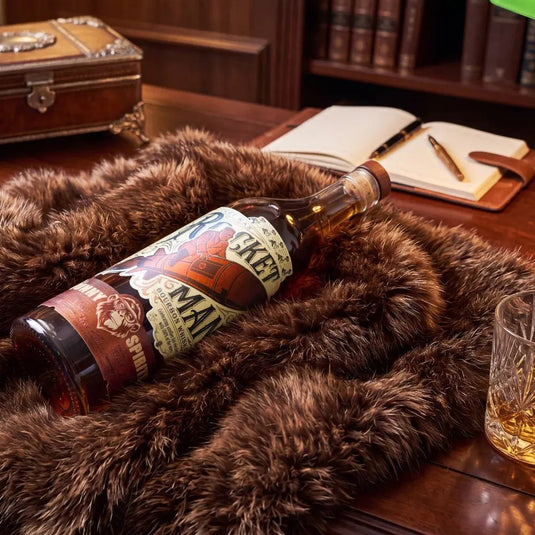 A bottle of Rocket Man Bourbon Whiskey, a limited release from Rocket Man, sits on a fur throw atop a wooden desk, accompanied by a whiskey glass, open notebook, pen, and books in the background.