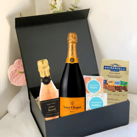 A black Premium Champagne Gift Box by Main Street Liquor sits open on a bed, featuring Veuve Clicquot champagne, Sugarfina bubbly bears, Ghirardelli chocolate squares, and a bath soak—a luxurious champagne gift for any special occasion.