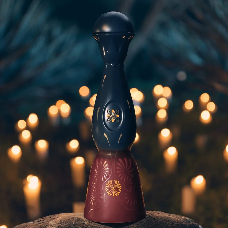 Load image into Gallery viewer, A dark blue and maroon Clase Azul Día de Muertos Trascender 2026 Limited Edition Añejo Tequila 1L decanter with floral engravings, set on a rock against a background of blurred glowing candles.
