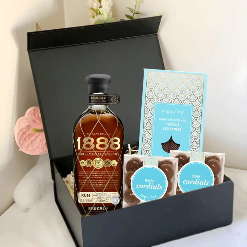 Load image into Gallery viewer, The Main Street Liquor Premium Rum Gift Box includes Brugal 1888, Sugarfina Rum Cordials, dark chocolate sea salted caramels, and two packs of rum cordial chocolates, beautifully arranged on a white surface with a white flower.
