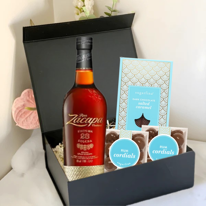 Load image into Gallery viewer, The Premium Rum Gift Box by Main Street Liquor features a bottle of Ron Zacapa, a Sugarfina dark chocolate sea salt caramel bar, and two boxes of Sugarfina Rum Cordials, all presented on white shredded paper with a flower in the background.
