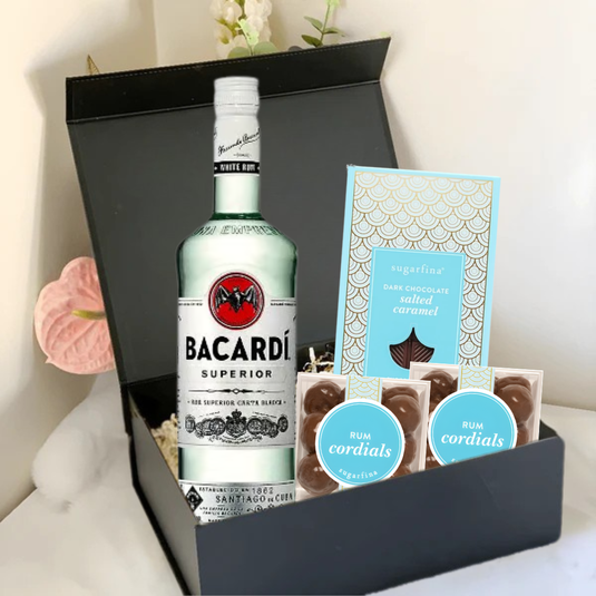 The Premium Rum Gift Box by Main Street Liquor, including Bacardi Superior, Sugarfina dark chocolate sea salt caramels, and two packs of Sugarfina Rum Cordials, is displayed on a white surface with pink flowers in the background.