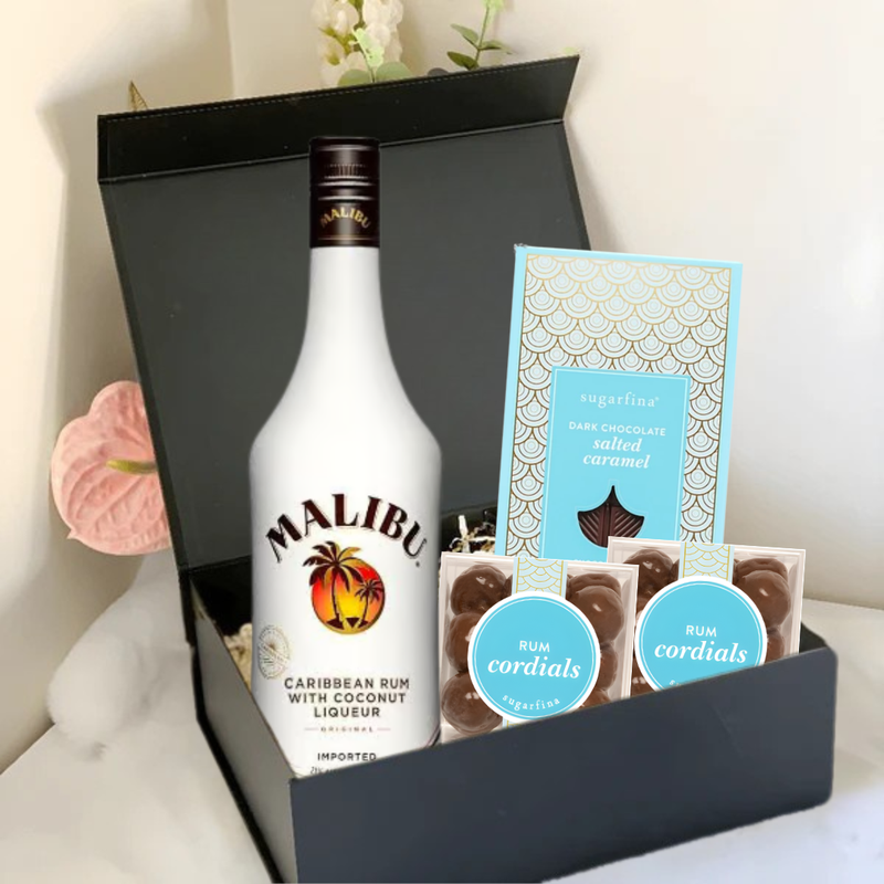 Load image into Gallery viewer, The Premium Rum Gift Box by Main Street Liquor features Malibu Caribbean Rum with Coconut Liqueur, Sugarfina dark chocolate sea salt caramels, and two packs of Sugarfina Rum Cordials. Pink flowers provide an elegant background touch.
