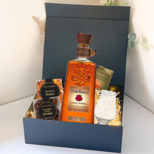 The Premium Bourbon Gift Box by Main Street Liquor includes Four Roses Single Barrel, two snack containers, a candle, caramel popcorn, a glass, and decorative shredded paper—beautifully arranged on a light surface.