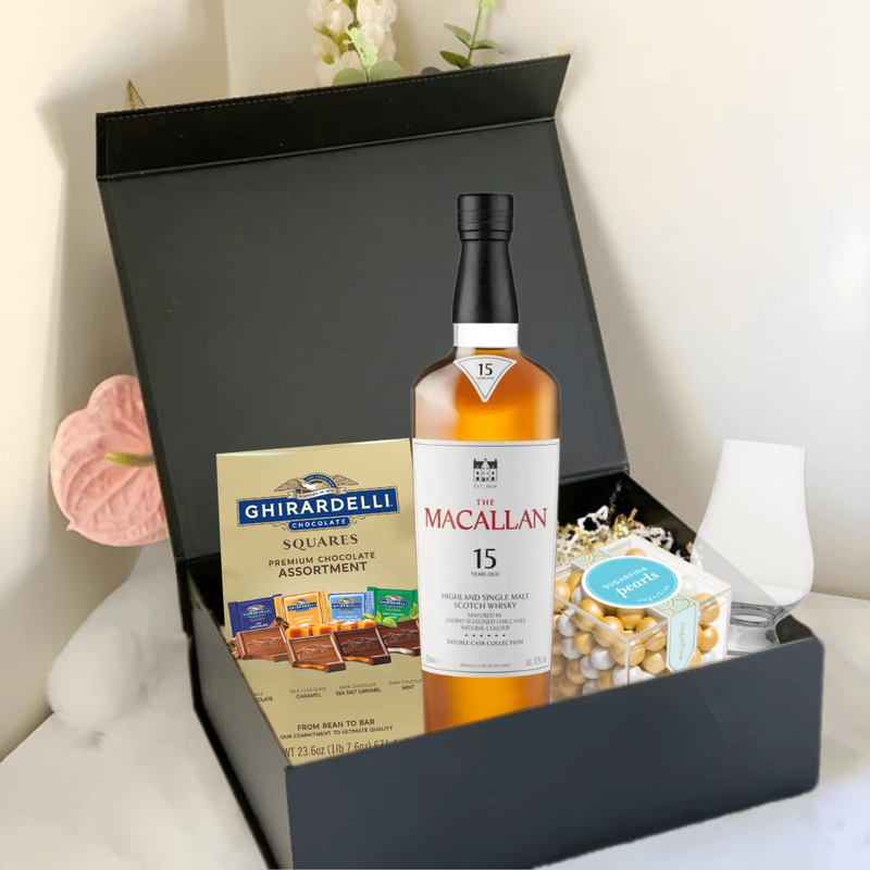 Load image into Gallery viewer, The Premium Scotch Gift Box by Main Street Liquor features The Macallan 15 Scotch, Ghirardelli chocolates, a glass, mixed nuts, and gold-wrapped treats—stylishly presented on white with pink flowers.
